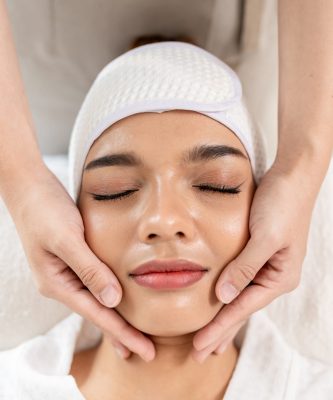 Beautiful woman getting head and facial massage treatment at beauty spa. Attractive girl lying down on massage bed, getting relaxing physiotherapy service from masseuse for body care in spa clinic.